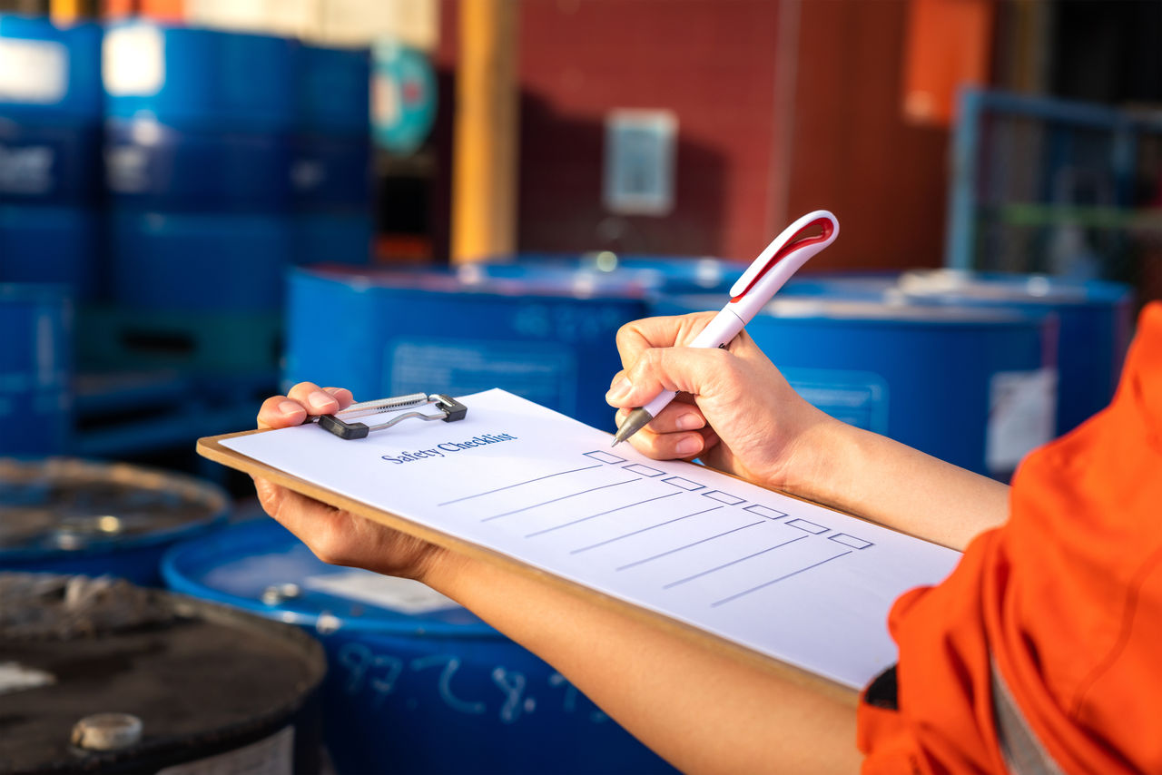 Safety inspection on the checklist document action with blurred background of chemical storage area at factory. Industrial safety concept photo. Selective focus at human's hand part., Safety inspection on the checklist document action with blurred 